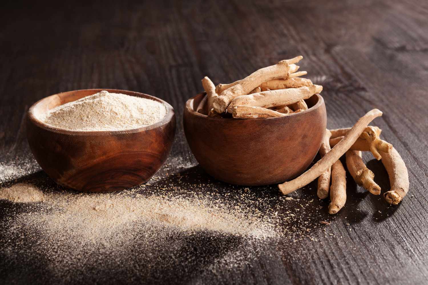 Dried ashwagandha roots in a wooden bowl beside milled ashwagandha root powder on a dark wood surface.